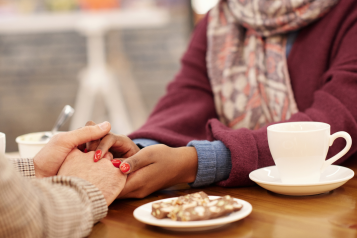 A photo of two women sat in a cafe, one holds the other's hands in hers over the table in support.